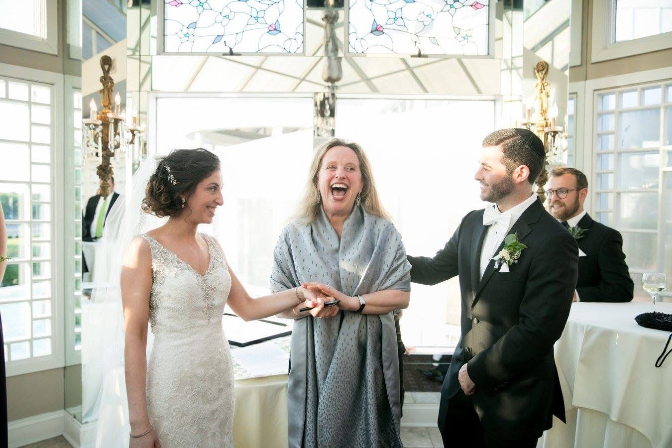 Rabbi Abby Sosland laughing as she joins a bride and groom’s hands at their wedding